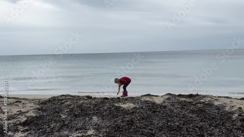 Blonde child girl in red nordic pants and shirt walks on empty sea beach. Kid picking up seashells and stones. Wide shot