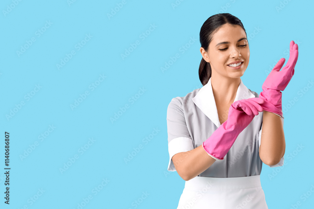 Young chambermaid putting on rubber gloves against blue background Young chambermaid putting on rubber gloves against blue background