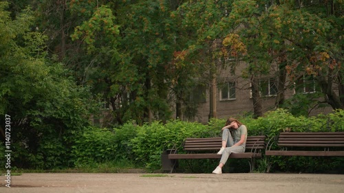 Young woman sits on wooden bench in park, resting head on arm in reflective, somber posture, surrounded by lush greenery and trees, expressing solitude and contemplation in peaceful outdoor setting photo