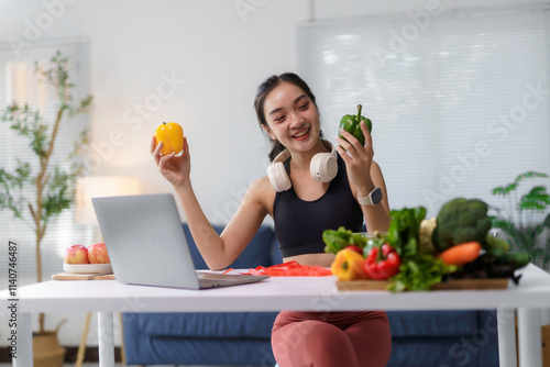 Smiling woman with headphones holding bell peppers, surrounded by fresh vegetables, using a laptop in a modern kitchen setting photo