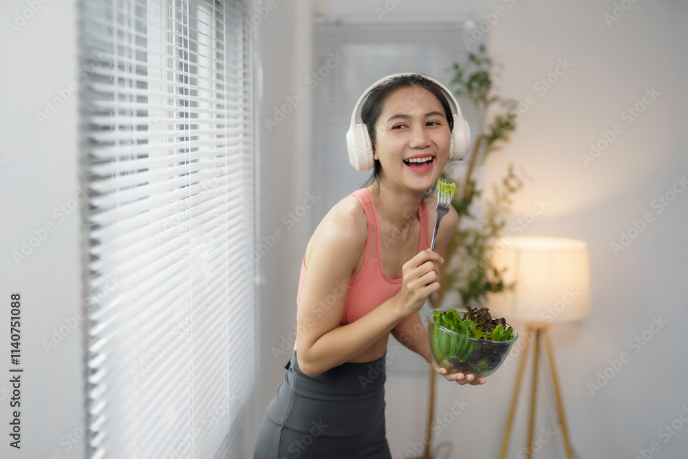 Asian woman wearing sportswear and headphones is happily eating a healthy salad at home