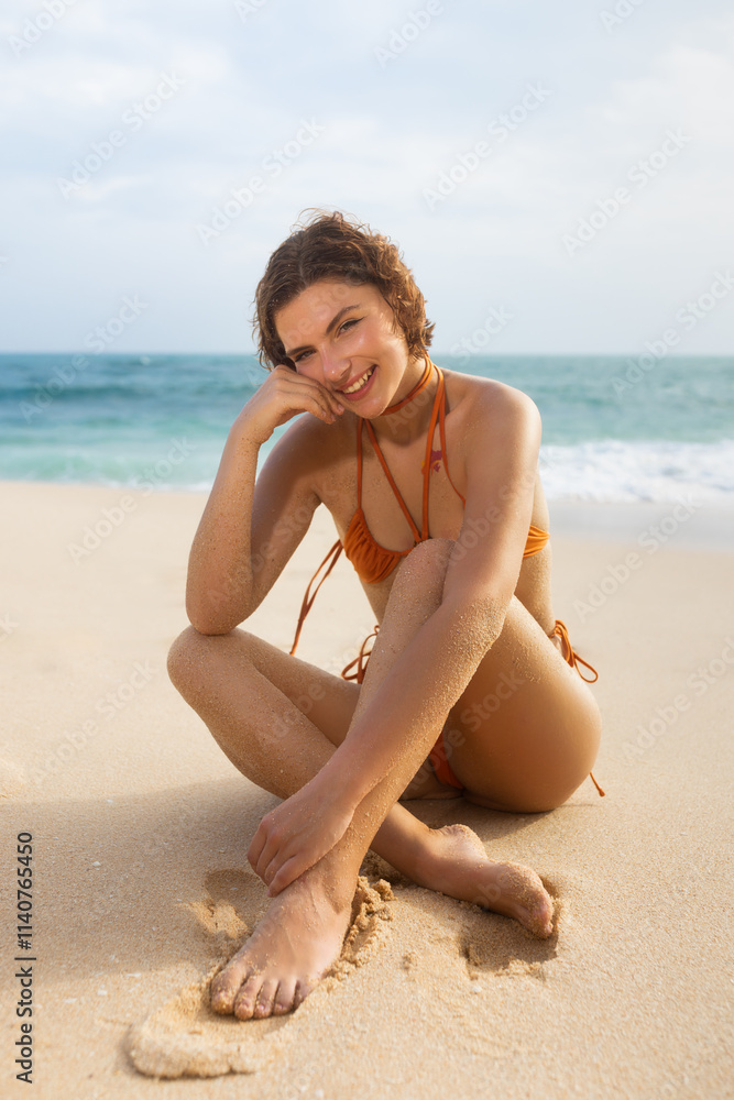 Smiling woman in an orange bikini sitting on the sandy beach with the ocean in the background. Perfect for summer, beach fashion, and lifestyle themes. Captures a joyful and relaxed moment. Smiling woman in an orange bikini sitting on the sandy beach with the ocean in the background. Perfect for summer, beach fashion, and lifestyle themes. Captures a joyful and relaxed moment.