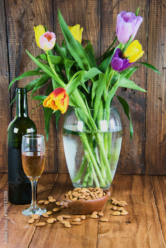 Tulip flowers in a transparent vase accompanied by a bottle of wine, glass and nuts on a rustic wooden table and background