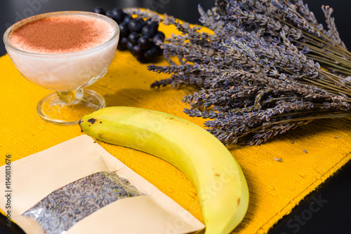 A vibrant arrangement of dessert ingredients on a yellow mat. A creamy dessert dusted with cocoa, fresh grapes, a banana, and dried lavender suggest a unique culinary creation. photo