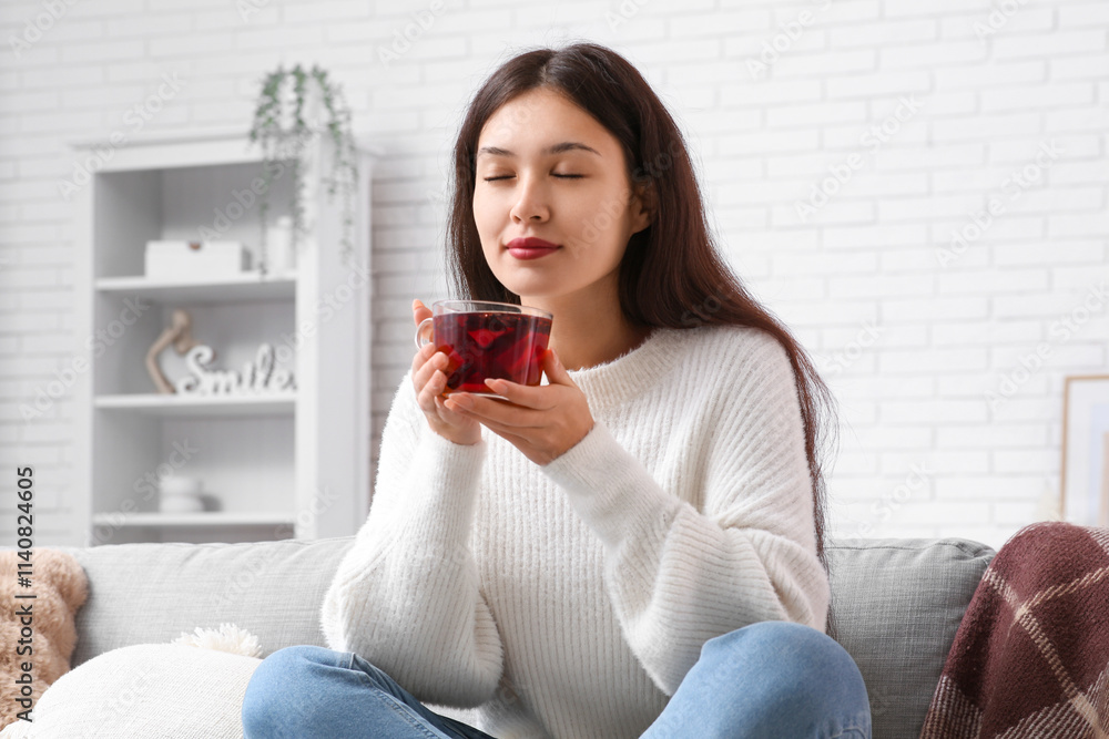 Beautiful young happy Asian woman with cup of hot mulled wine sitting on sofa at home Beautiful young happy Asian woman with cup of hot mulled wine sitting on sofa at home