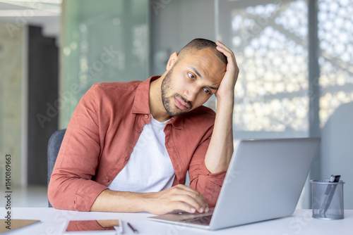Fotografie A man sitting at a desk in a modern office, appearing frustrated while using his laptop