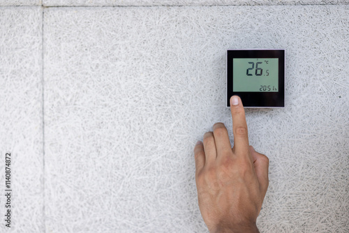 A person adjusts a digital thermostat mounted on a textured wall, displaying degrees Celsius. photo