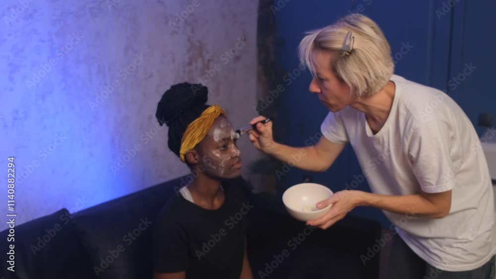 Two women are in a room. One woman with a yellow headscarf has a facial mask applied to her face with a brush by another woman. A white bowl is being held by the woman applying the mask Two women are in a room. One woman with a yellow headscarf has a facial mask applied to her face with a brush by another woman. A white bowl is being held by the woman applying the mask