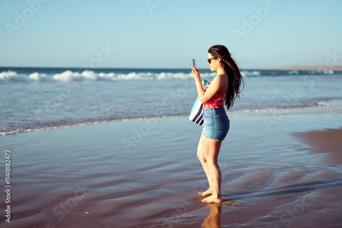 Latin woman taking photos with cell phone on the beach at sunset, vacations and summer