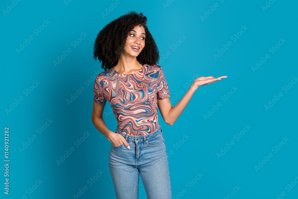 Young woman with curly hair poses stylishly against a vibrant blue background, wearing a patterned top and jeans