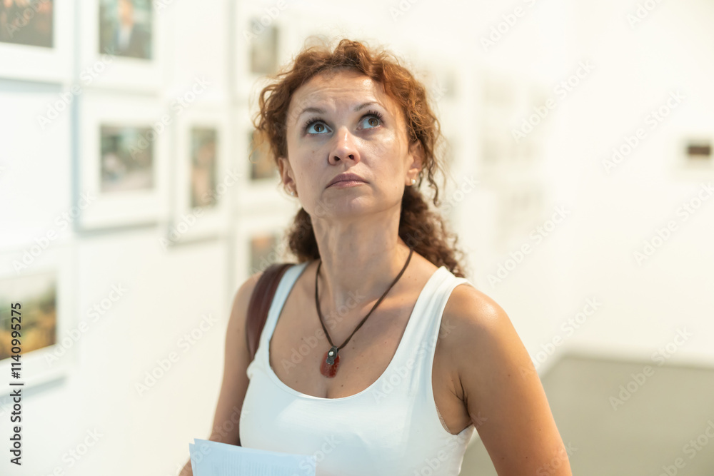 Woman in mid-forties, with curly auburn hair, observing framed photographs on display in modern photo gallery, showcasing deep interest in visual art Woman in mid-forties, with curly auburn hair, observing framed photographs on display in modern photo gallery, showcasing deep interest in visual art