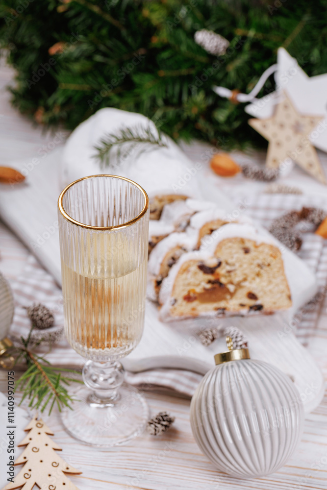 Naklejka premium A champagne glass with sparkling wine is displayed alongside stollen bread, almonds, pinecones, and decorative Christmas ornaments on a festive holiday-themed table.