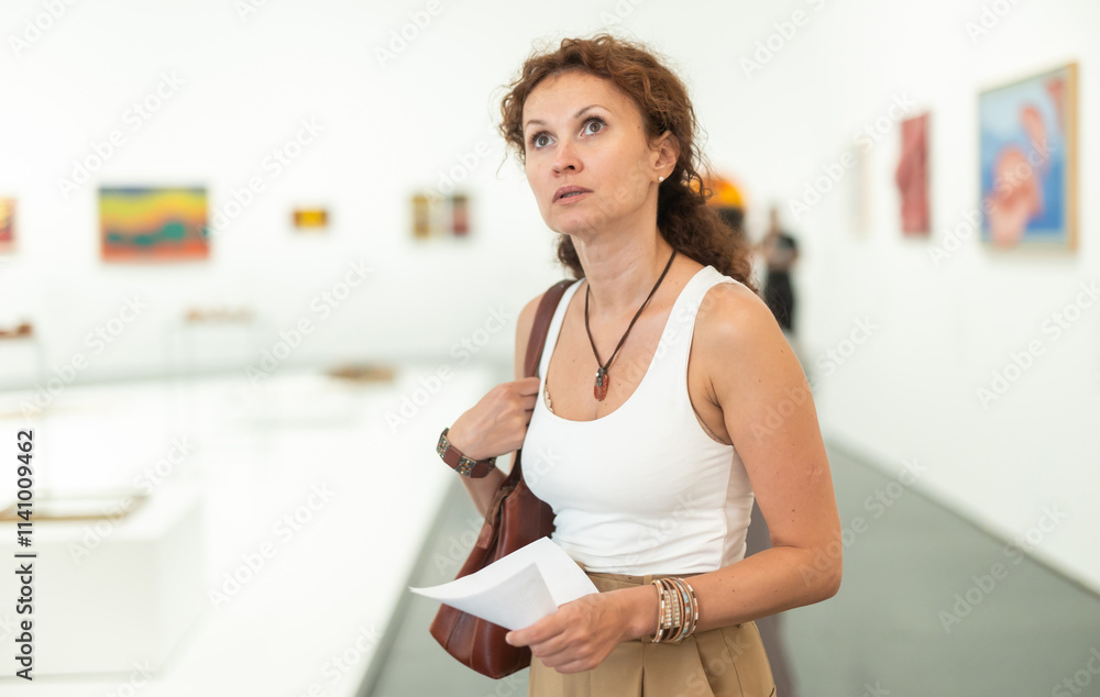 Interested middle-aged woman looking at different pictures placed on wall in museum Interested middle-aged woman looking at different pictures placed on wall in museum