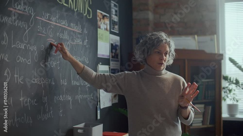 Mature female teacher with grey hair pointing at sentence written on blackboard and explaining English grammar rules during lesson in classroom