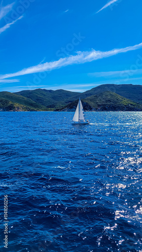 One sailboat with a white sail goes on the blue sea, lake or ocean. In the background are mountains covered with forests, and stone shore and a small town.