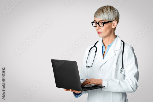 Modern doctor appointment concept. Woman doctor in white coat and glasses writing medical history in laptop isolated on light background, studio shot