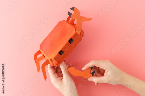 assembling details of upcycled egg box crab craft, creative process in progress, top view of hands, pink background, photo