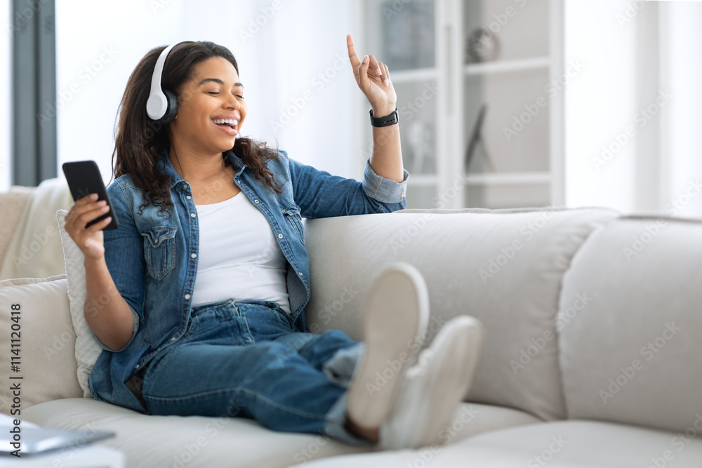 A young black woman sits comfortably on a couch, wearing headphones and smiling, as she listens to music on her phone. The warm daylight enhances the cozy atmosphere of her living space. A young black woman sits comfortably on a couch, wearing headphones and smiling, as she listens to music on her phone. The warm daylight enhances the cozy atmosphere of her living space.