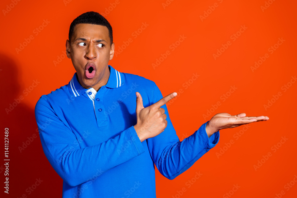 Stylish young man in blue shirt pointing with surprise on orange background Stylish young man in blue shirt pointing with surprise on orange background