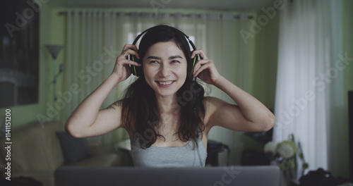 Portrait of young woman laughing sitting in front of computer at home. Female freelancer happily adjusting headset and put on glasses during remote work with laptop. Concept of lifestyle, 4k.