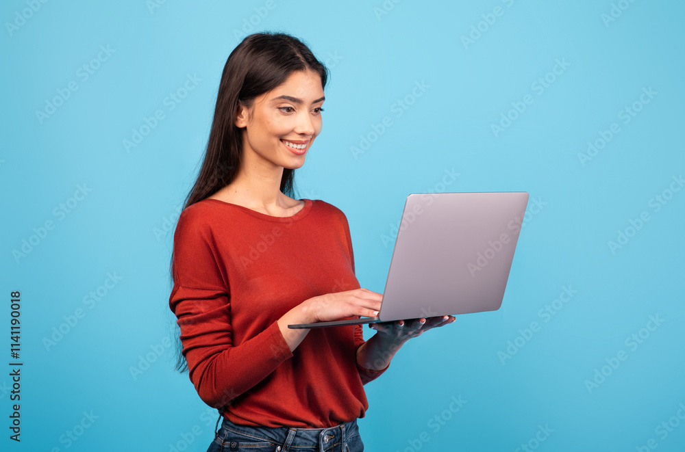 A young brunette woman stands in front of a vivid blue backdrop, happily typing on her laptop. She exhibits a relaxed and content demeanor, embodying a modern lifestyle. A young brunette woman stands in front of a vivid blue backdrop, happily typing on her laptop. She exhibits a relaxed and content demeanor, embodying a modern lifestyle.