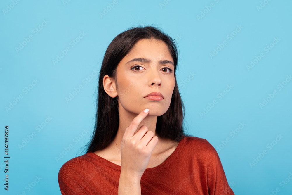 A millennial woman with brunette hair appears deep in thought. She gently touches her chin, conveying contemplation. The bright blue backdrop enhances her expression and lifestyle vibe. A millennial woman with brunette hair appears deep in thought. She gently touches her chin, conveying contemplation. The bright blue backdrop enhances her expression and lifestyle vibe.