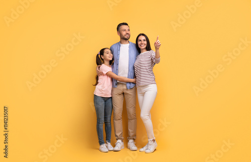 A European family stands together in front of a yellow backdrop, with the daughter hugging her father. The mother points out something, reflecting joy and connection among them.