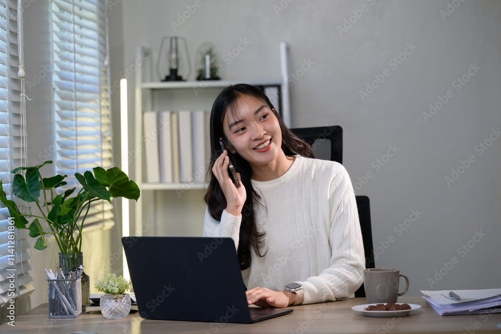 Young asian woman working on a laptop while chatting on the phone, sitting at her desk in a cozy home office, experiencing a fulfilling and productive workday filled with focus and creativity