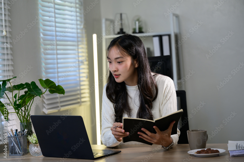 Young asian businesswoman is taking notes in her notebook while attending an online meeting on her laptop in her brightly lit home office Young asian businesswoman is taking notes in her notebook while attending an online meeting on her laptop in her brightly lit home office