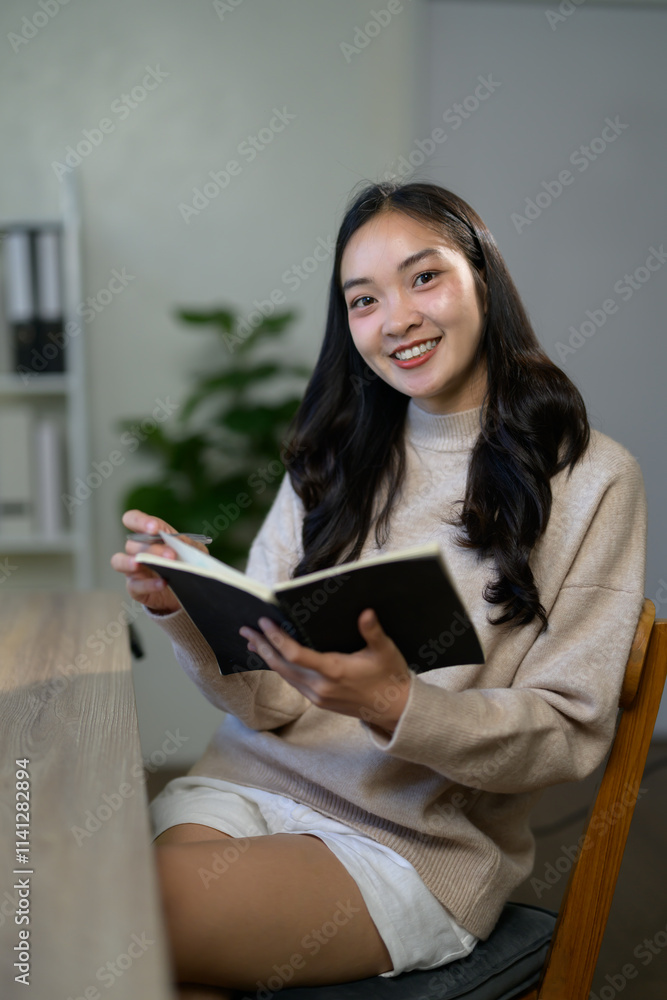 Happy young asian woman sitting at a wooden desk, holding a pen and notebook, smiling while taking notes and engaging in work or study from the comfort of her home office Happy young asian woman sitting at a wooden desk, holding a pen and notebook, smiling while taking notes and engaging in work or study from the comfort of her home office