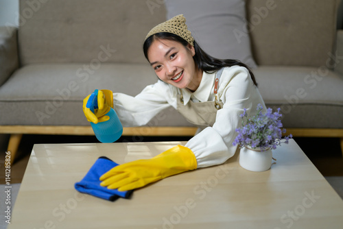 Young asian housekeeper wearing yellow gloves and apron, cleaning living room table with microfiber cloth and disinfectant spray bottle, smiling towards camera photo