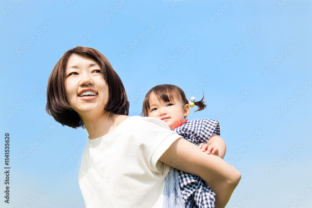 青空を背景に女の子の幼児を背負い育児をする若いお母さん。育児,家族,愛情のイメージ