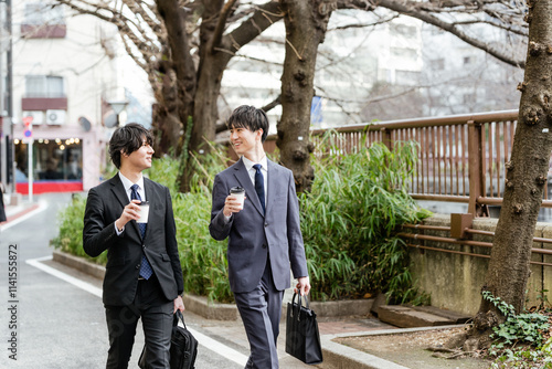 会話しながらコーヒーを手に持って顔を見合わせ並んで歩く2人のスーツの男性 photo