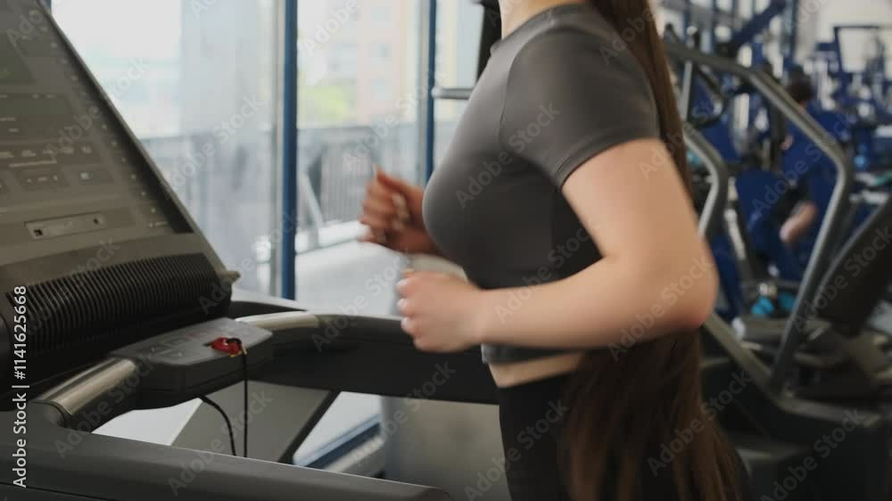 Sportswoman runs on treadmill looking at monitor in spacious gym. Young woman in sportswear develops calves and thighs increasing endurance Sportswoman runs on treadmill looking at monitor in spacious gym. Young woman in sportswear develops calves and thighs increasing endurance