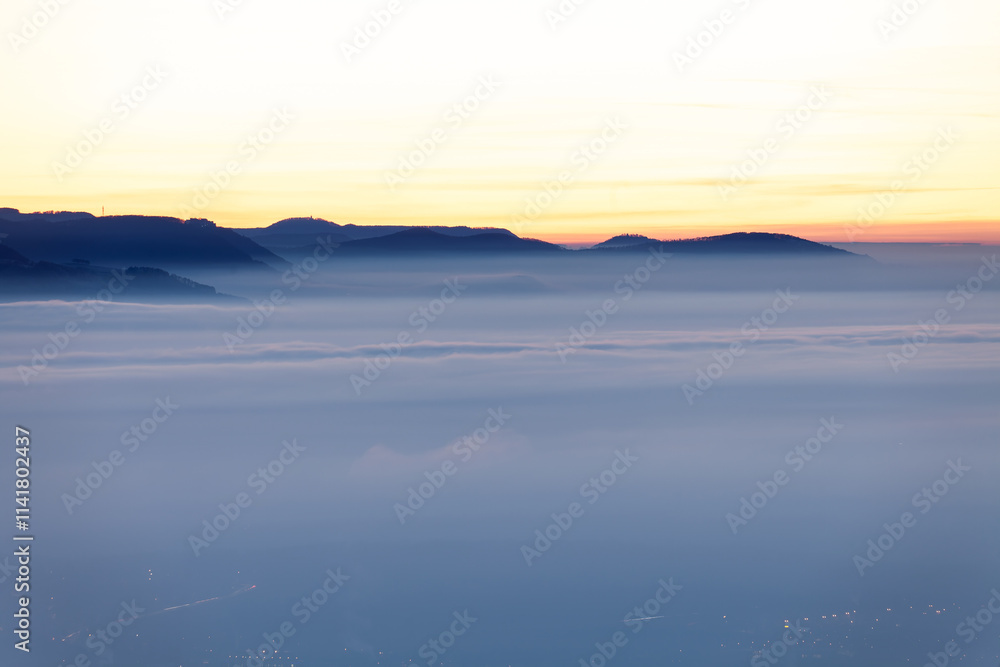 Fototapeta premium Ausblick auf das Nebelmeer im Tal. Am Horizont die Burg Hohenneuffen und die Achalm bei Reutlingen. Schwäbische Alb Albtrauf Silhouetten im Winter zur Blauen Stunde.