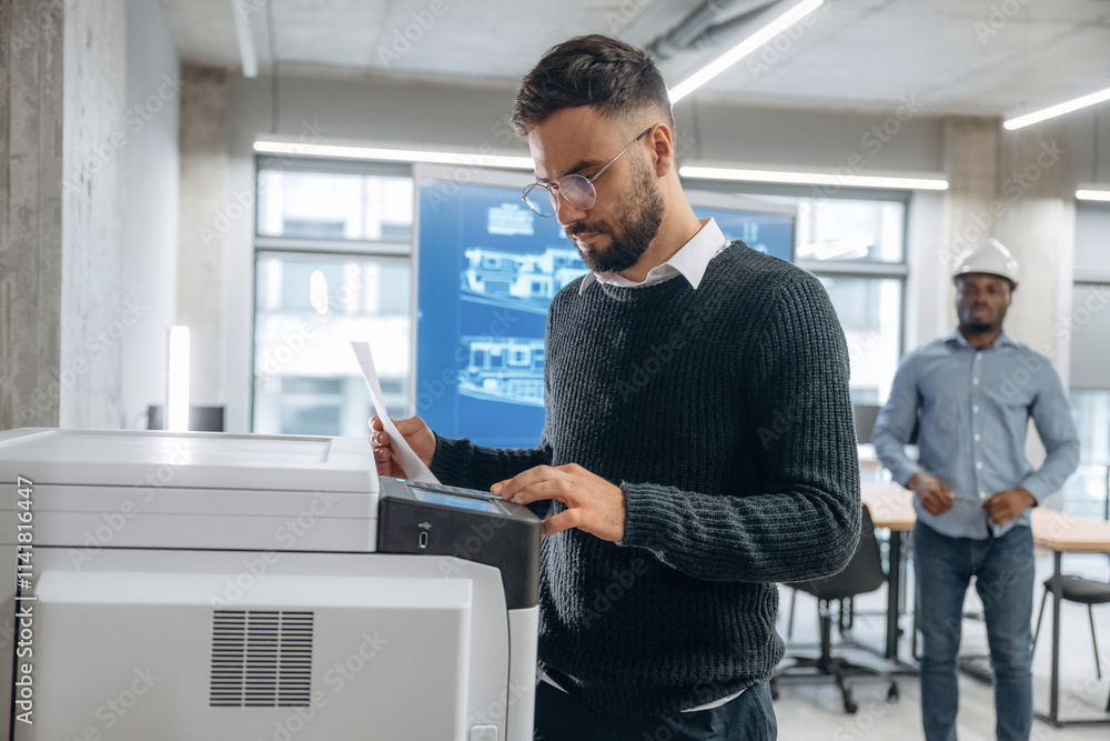 Two office workers are standing, man is with documents near printer Two office workers are standing, man is with documents near printer