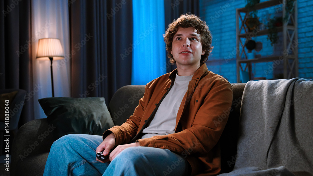 Young man spending evening at home sitting on sofa in living room, watching tv, evening with movie or news program, positive smiling expression. Young man spending evening at home sitting on sofa in living room, watching tv, evening with movie or news program, positive smiling expression.