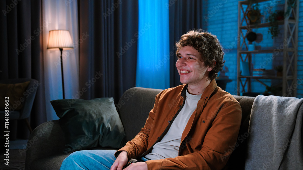 Young man spending evening at home sitting on sofa in living room, watching tv, evening with movie or news program, positive smiling expression. Young man spending evening at home sitting on sofa in living room, watching tv, evening with movie or news program, positive smiling expression.