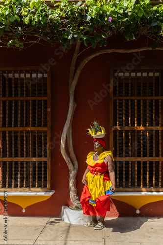 Woman In A Colorful Dress With A Fruit Basket On Her Head Standing Outside A Colonial Building With Wooden Windows