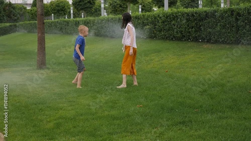 Children are running on wet green grass with automatic irrigation turned on in the city park, hot summer weather. Children have fun in the summer playing with automatic lawn watering.