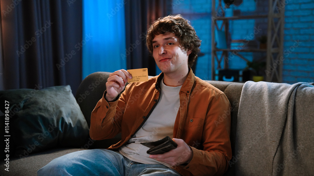 Fototapeta premium Young man spending evening at home sitting on sofa in living room, holding gold credit card in hand looking happy, smiling positive expression.
