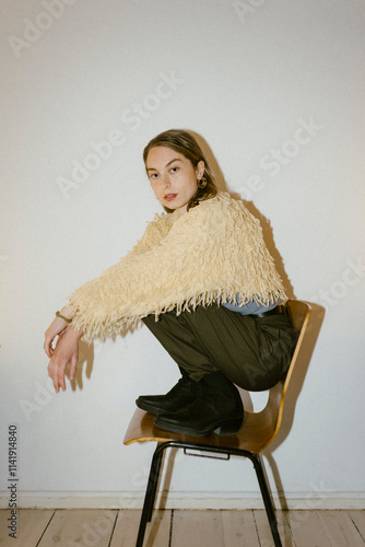 Portrait of fashionable young woman wearing fake fur jacket crouching on chair near wall at home photo