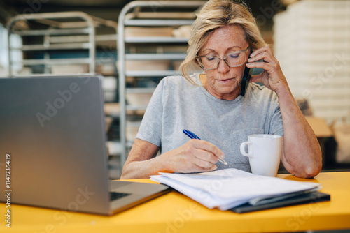 Senior female baker talking on smart phone while preparing notes at bakery photo