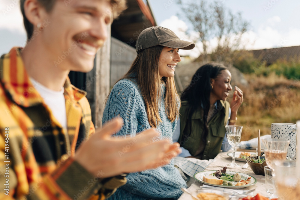 Smiling young woman wearing cap sitting with male and female friends at dinner party during social event Smiling young woman wearing cap sitting with male and female friends at dinner party during social event