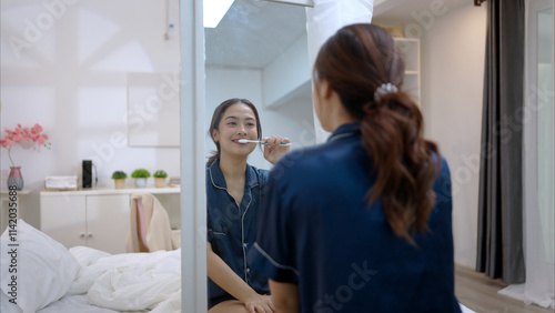 Asian young woman in a blue pajama is brushing her teeth in front of the mirror before taking a shower