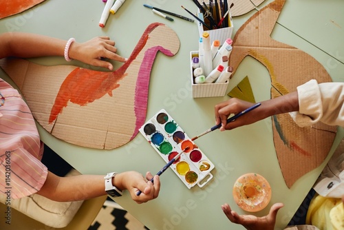 Top down view at group of children painting cardboard dinosaur figures using colorful watercolors during art and craft class copy space