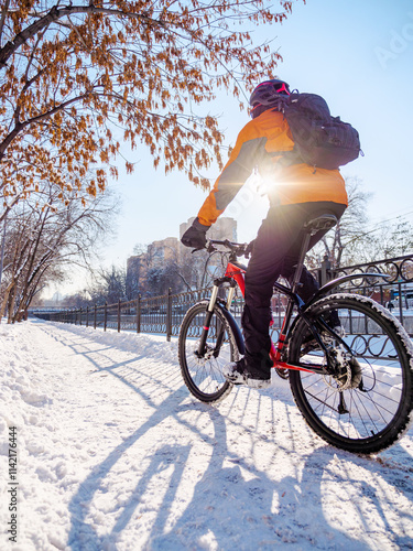 A bicyclist rides through a winter city on a sunny day. A guy in a bicycle helmet and orange jacket rides a red bicycle. Ecological transportation