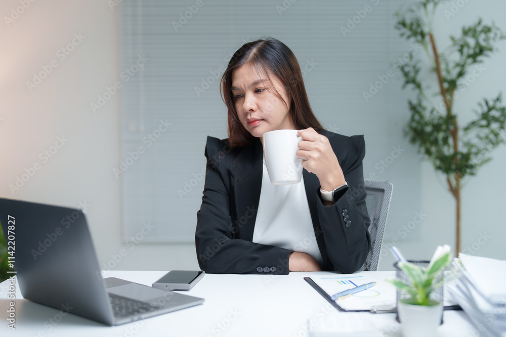 Asian businesswoman drinking coffee while working with laptop and documents at her desk in office, she is wearing a black suit and a white shirt Asian businesswoman drinking coffee while working with laptop and documents at her desk in office, she is wearing a black suit and a white shirt