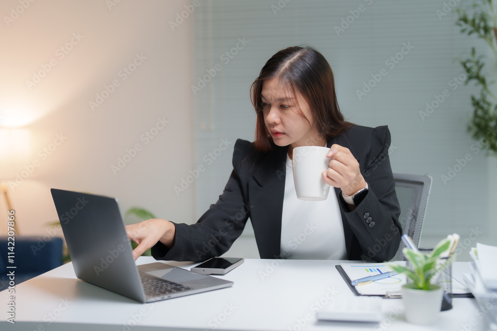 Young Asian businesswoman sipping coffee while working on a laptop at her modern office desk, analyzing financial documents and planning business projects Young Asian businesswoman sipping coffee while working on a laptop at her modern office desk, analyzing financial documents and planning business projects