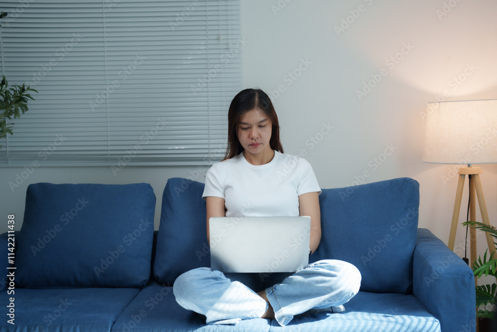 Young Asian woman sitting cross-legged on a comfortable blue sofa, using a laptop at home, working remotely and browsing the internet in a cozy living room filled with natural light Young Asian woman sitting cross-legged on a comfortable blue sofa, using a laptop at home, working remotely and browsing the internet in a cozy living room filled with natural light